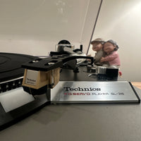 Technics turntable with a needle on a gray surface, with a blurred background of two people and a lamp.